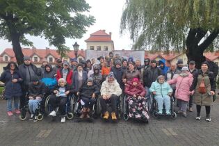 Grupa osób pozuje ustawiona w kilku rzędach do zdjęcia na tle budynków o czerwonych dachach. Zdjęcie wykonane na świeżym powietrzu w pochmurny dzień.