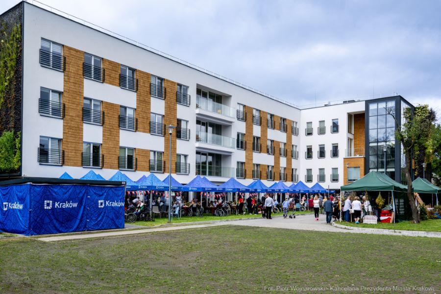 Na zdjęciu namioty ze stoiskami oraz namioty Miasta Krakowa, gdzie siedzą mieszkańcy oraz zaproszeni mieszkańcy z innych DPS-ów w ogrodzie na tle Domu. 