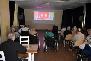 Na zdjęciu widać grupę starszych osób siedzących na krzesłach i w wózkach inwalidzkich w ciemnej sali, oglądających film wyświetlany na dużym ekranie. 