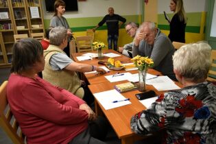 Kilka osób starszych siedzi przy stole, na którym leżą kartki papieru, długopisy i książki. Dwie kobiety stoją i prowadzą zajęcia. Na stole stoją wazony z żółtymi kwiatami.