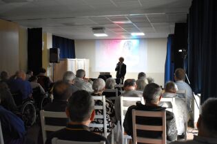 W sali z rzędem krzeseł i wózków inwalidzkich siedzi grupa starszych osób, oglądających występ mężczyzny z mikrofonem na tle ekranu z kolorową projekcją.