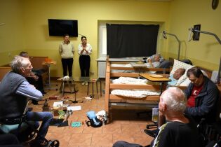 W pokoju dwie kobiety stoją przy ścianie i prowadzą występ dla grupy starszych osób, z których część leży w łóżkach lub siedzi na wózkach inwalidzkich. Na podłodze leżą różne rekwizyty. 