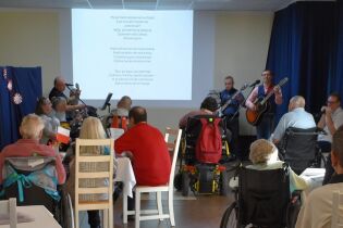 Grupa osób, głównie seniorów, uczestniczy w koncercie z udziałem muzyków grających na gitarach. Na ekranie za nimi wyświetlany jest tekst piosenki. Widzowie, niektórzy na wózkach, śpiewają razem.