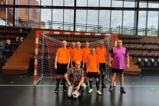 Drużyna w sportowych koszulkach pozuje do wspólnego zdjęcia przed bramką na hali sportowej. Obok zawodników stoi bramkarz i trener.