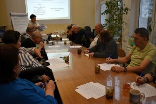 Grupa osób siedzi przy dużym stole w sali zajęciowej. Uczestnicy wykonują ćwiczenia pisemne, trzymając długopisy i kartki. Na końcu stołu stoi prowadząca, która omawia materiał.