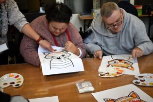 Dwie osoby siedzą przy stole i malują obrazki przedstawiające zwierzę – prawdopodobnie lisa. Po lewej stronie osoba trzyma kartkę i skupia się na malowaniu, a obok niej ktoś pomaga, wskazując miejsce.