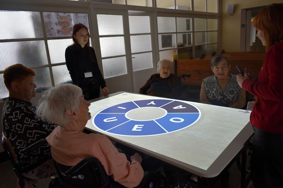 Kilka starszych osób siedzi wokół dużego stołu multimedialnego, na którego powierzchni wyświetlony jest jasny ekran z okrągłym diagramem zawierającym litery.