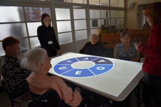 Kilka starszych osób siedzi wokół dużego stołu multimedialnego, na którego powierzchni wyświetlony jest jasny ekran z okrągłym diagramem zawierającym litery.