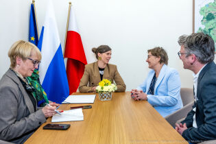W gabinecie przy jasnym prostokątnym stole przy którym siedzi 5 osób u szczytu stołu dwie uśmiechnięte kobiety rozmawiają ze sobą.   